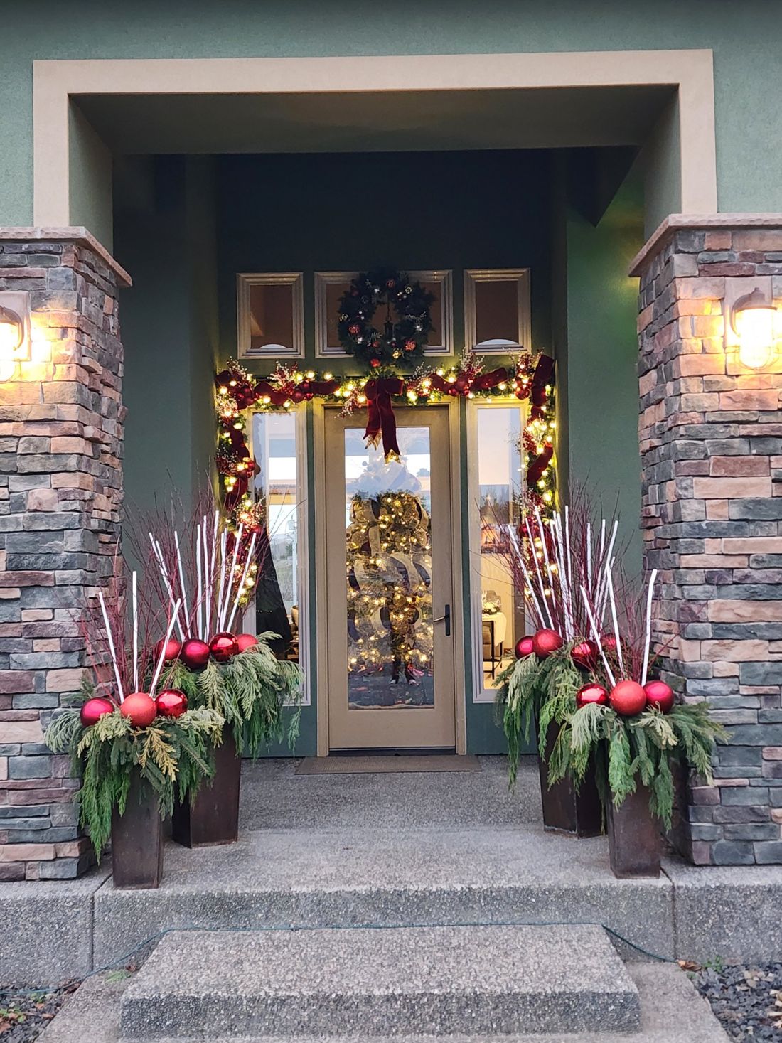 Front entry framed in lit garland with red bows and planters of greens, birch and red ornaments